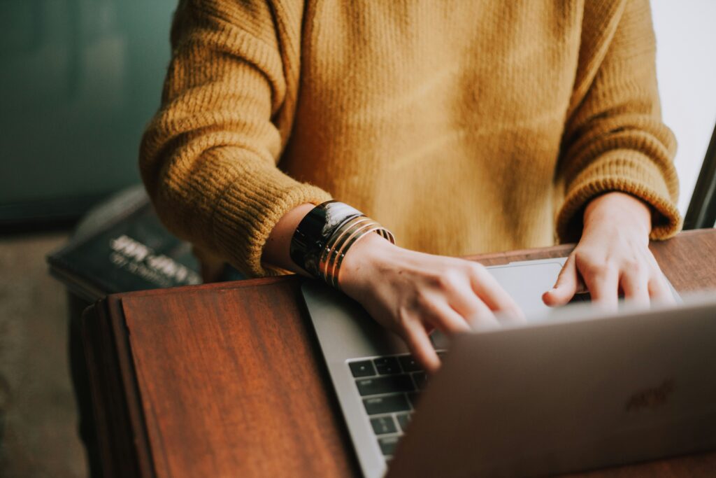 Woman in yellow shirt with gold bracelet sitting at table typing on a laptop. We cannot see her face.